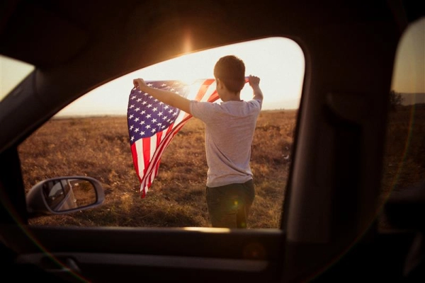 A young boy holds an American flag against a sunset, viewed through the passenger side of a car window