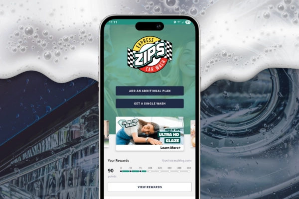 Phone displaying the home page of the ZIPS Car Wash app in front of a car headlight with soapy suds